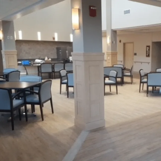 Community dining room with a serving counter, wood-look flooring and tables set with upholstered chairs