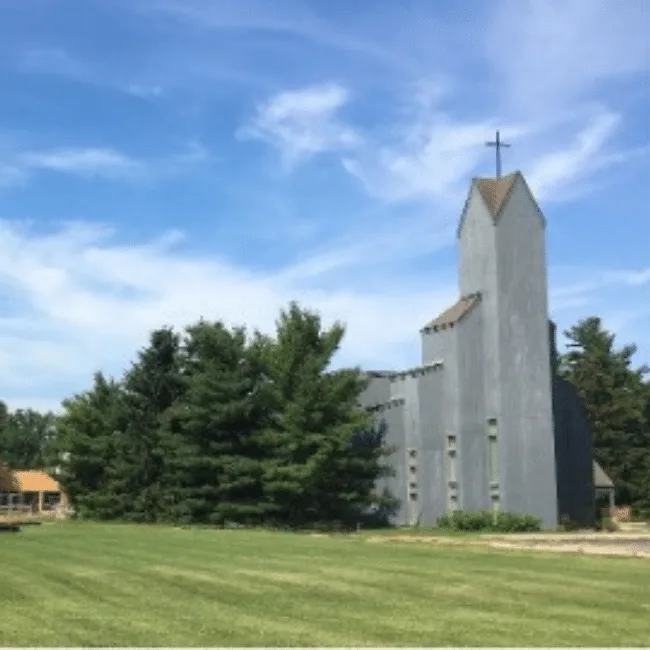 Church with a tall concrete bell tower topped by a cross, set behind mature evergreens